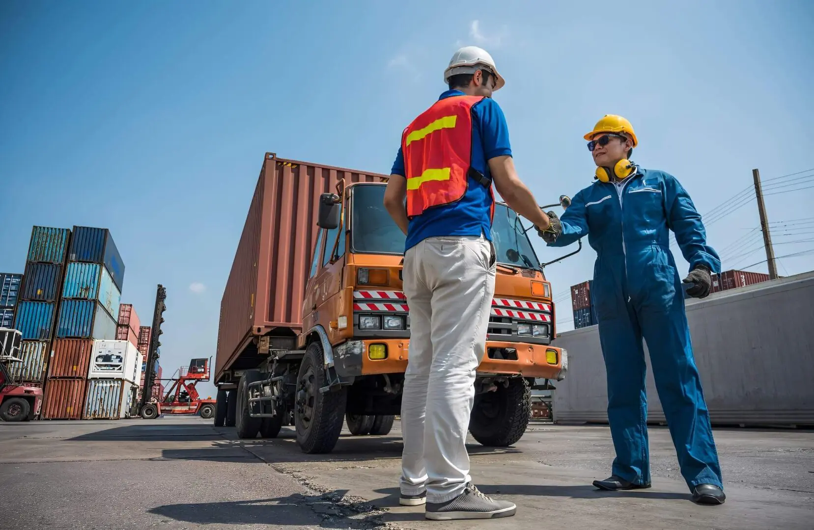 Two workers in safety gear discussing near a container truck at a shipping yard.