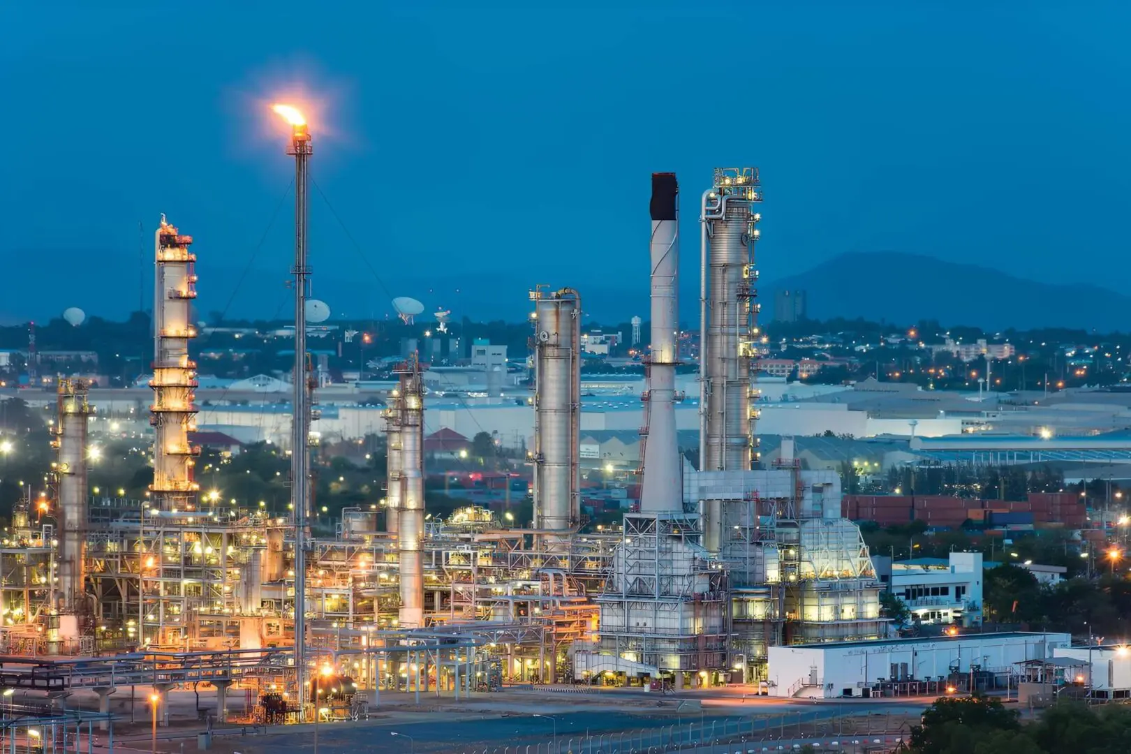 Industrial refinery complex illuminated at dusk with city lights in the background.