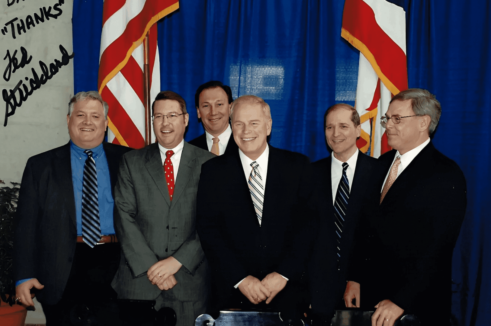 Six men in suits, smiling, with flags.